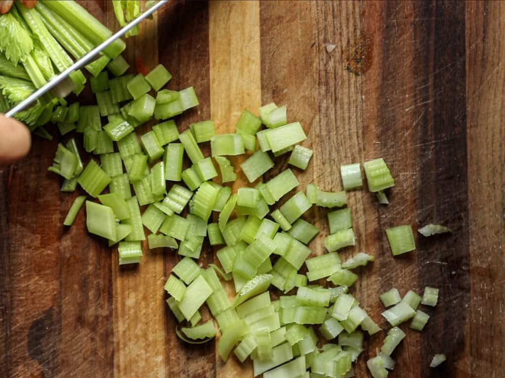 Chopped celery pieces scattered on a wooden cutting board, with a person’s hand holding a knife, continuing to slice celery stalks.